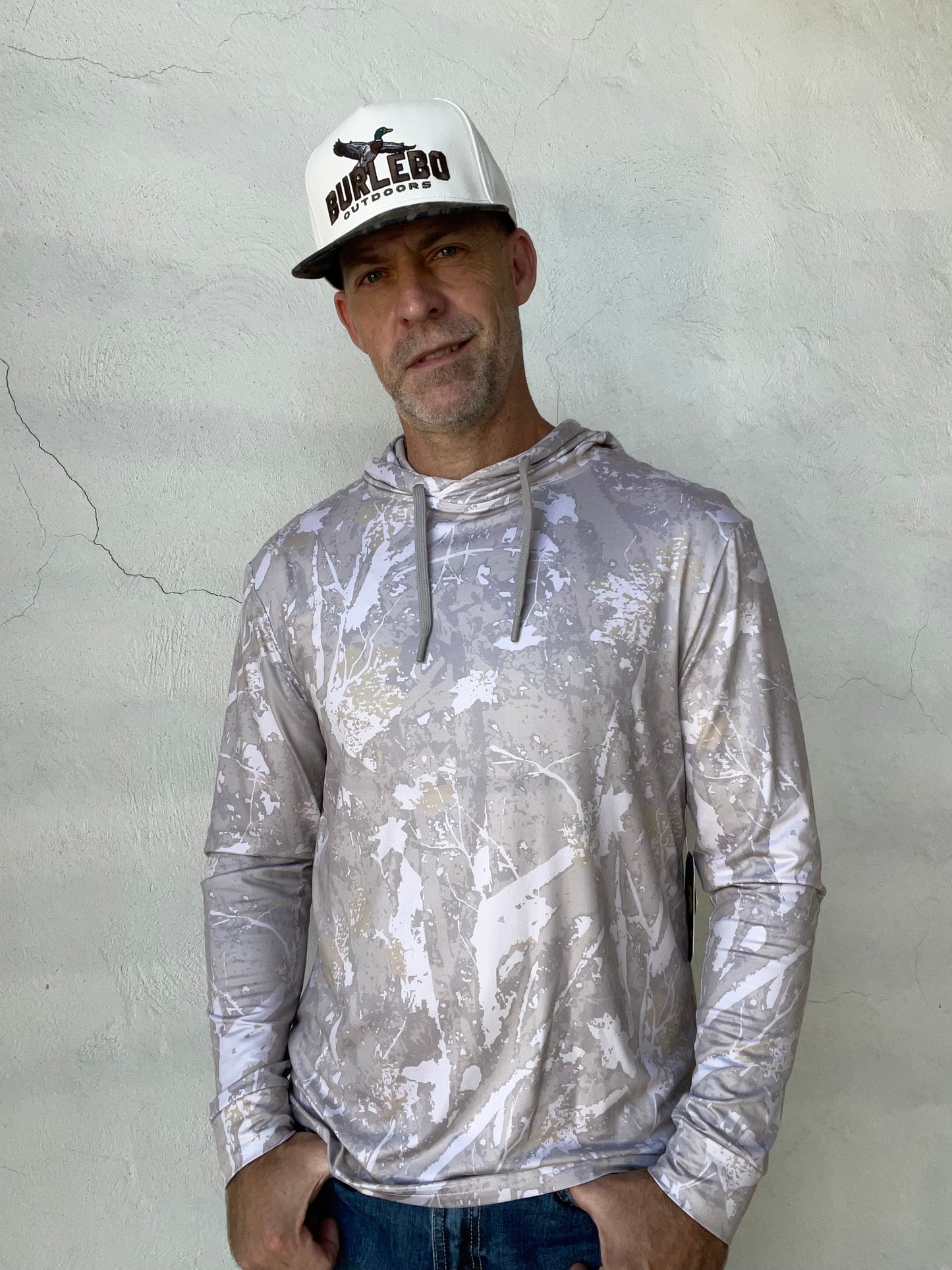 A man wearing a long-sleeve hooded shirt with a distressed silver and white paint splatter pattern, paired with a white 'Bulldog' trucker hat, standing against a textured light gray stucco wall.