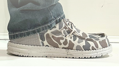 A pair of canvas lace-up shoes with a gray, beige, and brown giraffe-like camouflage pattern, shown with a brand tag and a faint reflection of a person wearing them at the bottom.