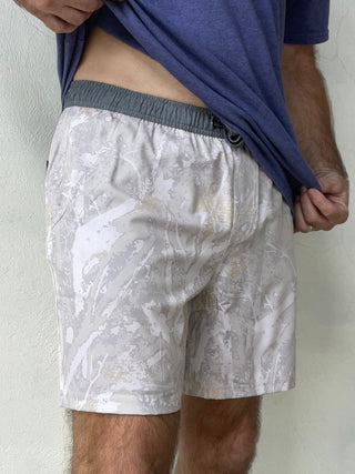 A man wearing light gray and white camouflage swim trunks with a dark gray elastic waistband, lifting his blue t-shirt to show the fit of the shorts.