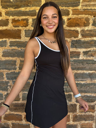 A young woman with long dark hair smiling in front of a rustic brick wall, wearing a black sleeveless tank dress with white trim.