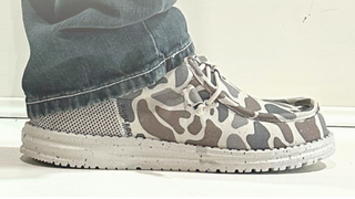A pair of canvas lace-up shoes with a gray, beige, and brown giraffe-like camouflage pattern, shown with a brand tag and a faint reflection of a person wearing them at the bottom.