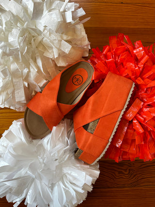 A pair of bright orange platform slide sandals with crisscross straps, styled on a wooden surface with white and red pom-poms.