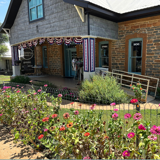 A historic stone and wood building with a front porch, surrounded by a landscaped garden with pink and red flowers in the foreground.