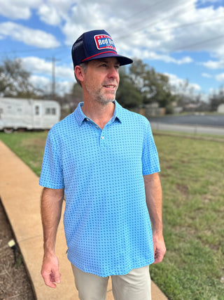Man wearing a blue patterned shirt and cap with a logo, standing outdoors on a sidewalk.