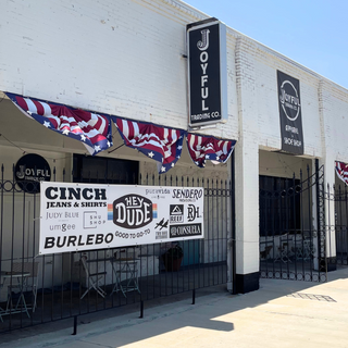 A storefront exterior of a white brick building with black metal gates, patriotic bunting, and multiple vertical signs including Joyful Trading Co. and a sign with the brands the store carries. A sidewalk runs along the front under a clear blue sky.