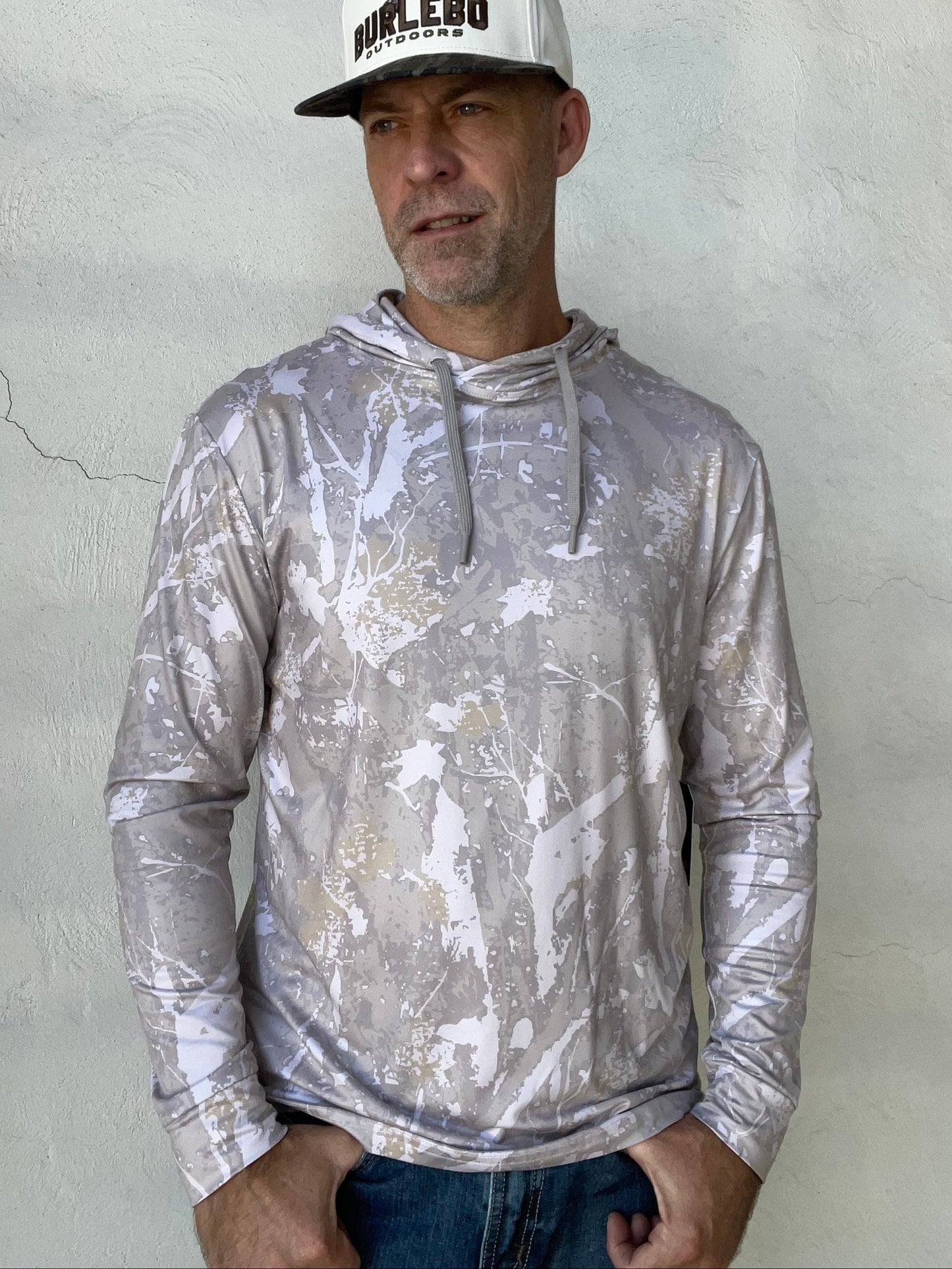 A man wearing a long-sleeve hooded shirt with a silver, white, and gray abstract splatter pattern, paired with a white and black trucker hat that reads 'DIELEBO'. He is standing against a textured, light gray stucco wall.