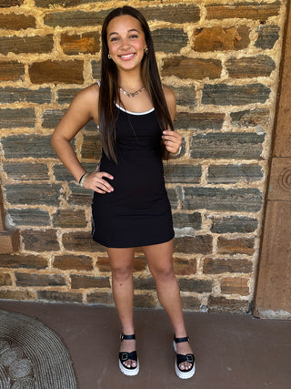 A young woman smiling in front of a rustic brick wall, wearing a black sleeveless mini dress with white trim and black platform sandals with white soles.