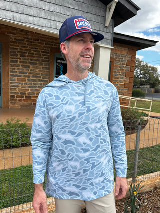 Man wearing a blue patterned shirt and cap standing outside a house.