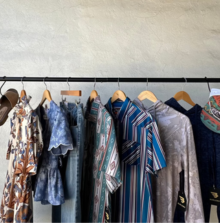 A clothing rack displaying a collection of diverse garments, including patterned dresses, striped shirts, and denim, hanging against a textured beige plaster wall.