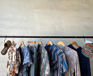 A clothing rack displaying a collection of diverse garments, including patterned dresses, striped shirts, and denim, hanging against a textured beige plaster wall.