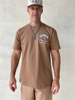 Man wearing a brown t-shirt with a logo and text, standing against a plain white wall.