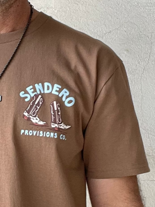 Man wearing a brown t-shirt with a logo and text, standing against a plain white wall.