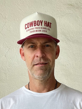 Man wearing a white t-shirt with red text and a white cap with red text against a beige wall.