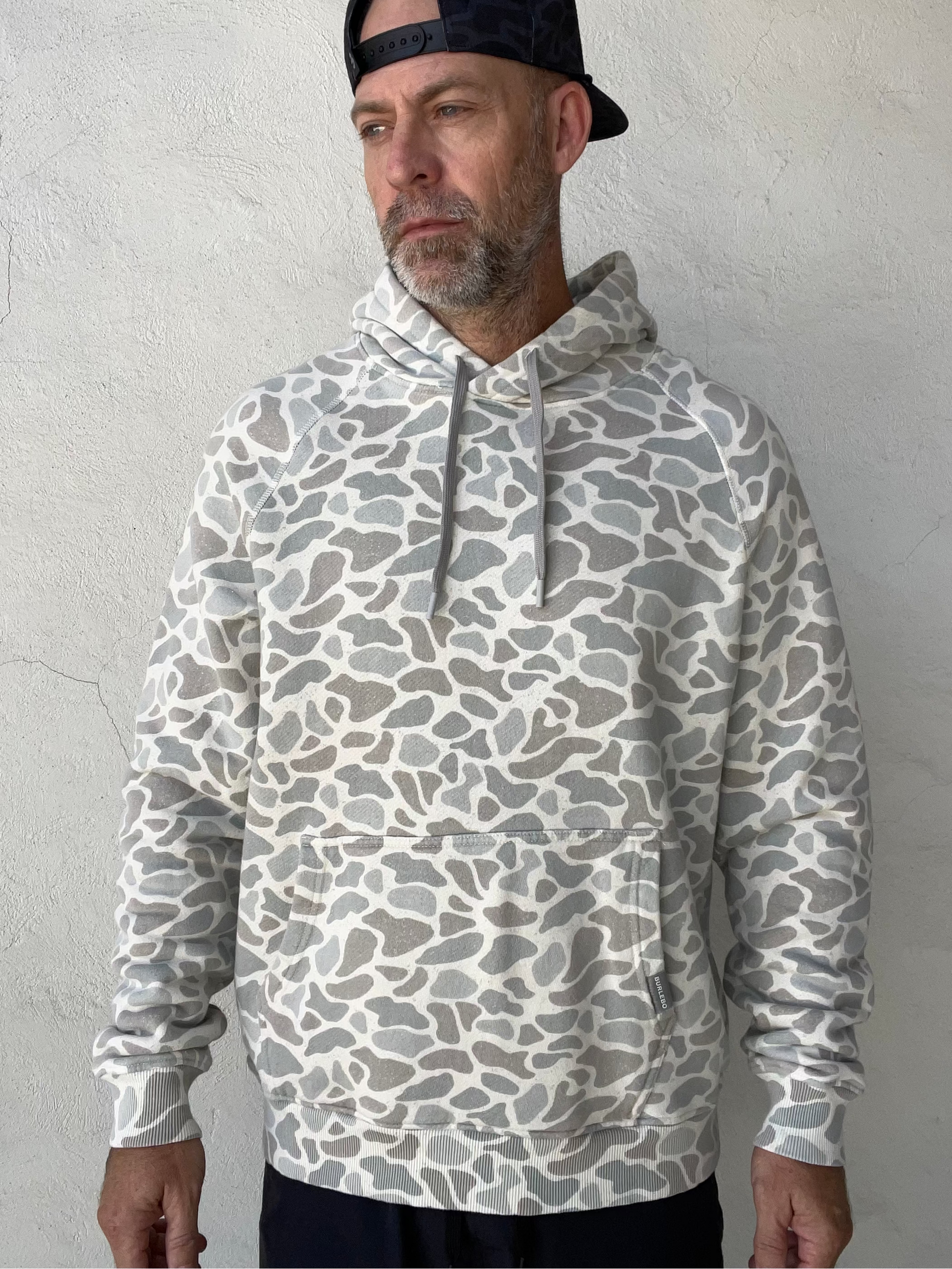 A man wearing a gray and white abstract patterned hoodie with a front kangaroo pocket and a black baseball cap, standing against a textured off-white wall.