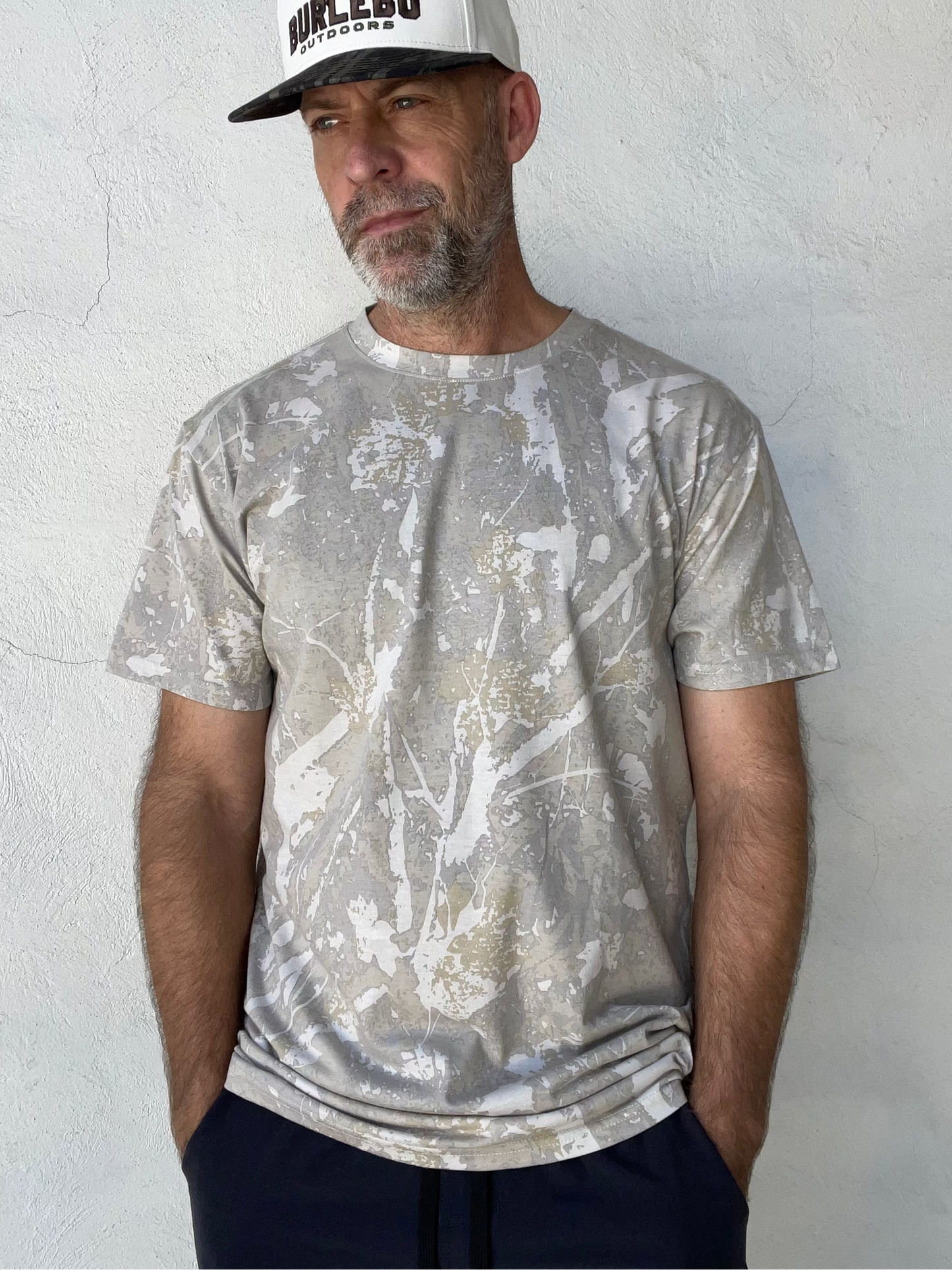 A man wearing a light gray and white splatter-patterned t-shirt and a white trucker hat with a black logo, standing against a textured white wall.