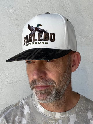 A man wearing a white and black snapback cap with a duck logo and 'BURLEBO OUTDOORS' text, paired with a distressed gray t-shirt, standing against a textured light gray wall.