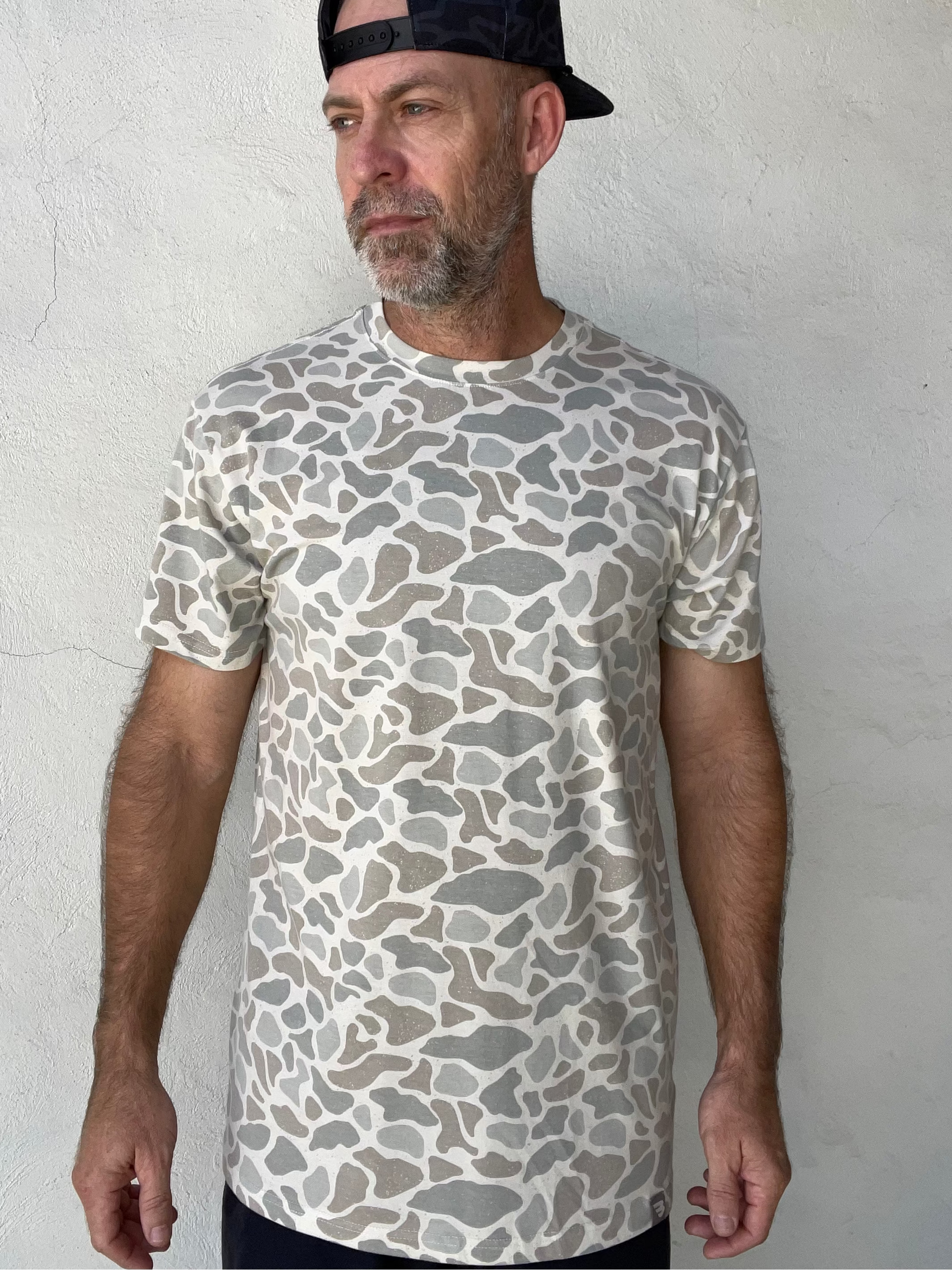 A man wearing a short-sleeve crewneck t-shirt with a gray and off-white organic stone-like pattern, paired with a dark patterned baseball cap, standing against a textured white stucco wall.