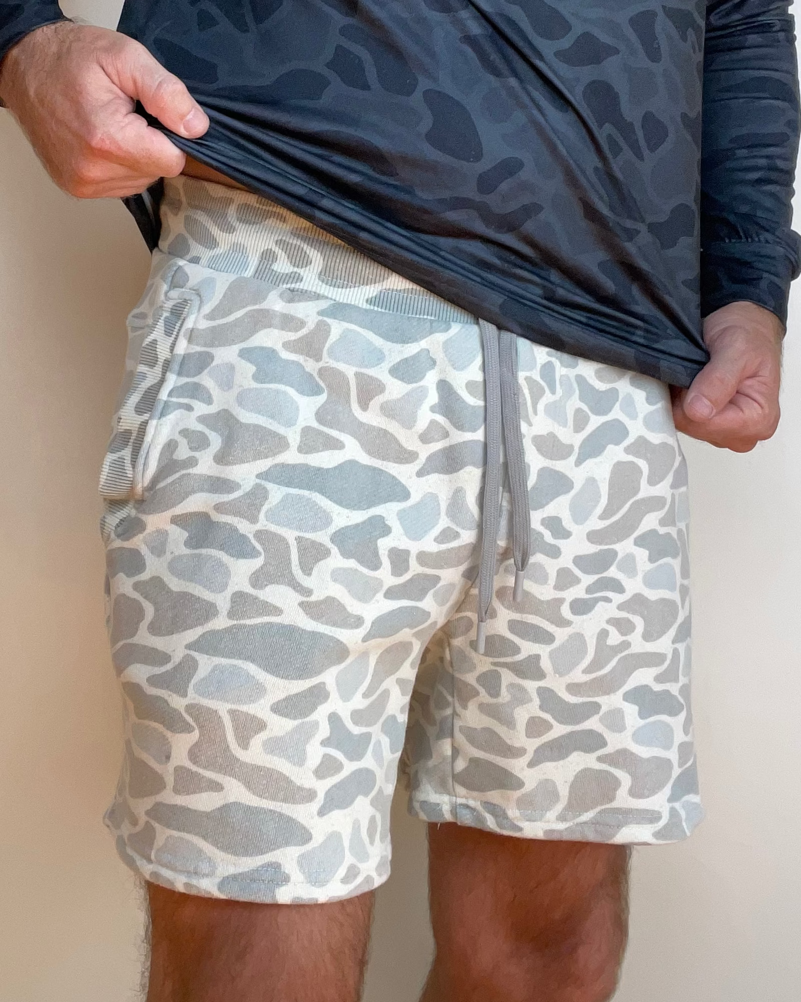 A man wearing a dark gray camouflage long-sleeve hooded shirt and light gray and white camouflage shorts, holding up the shirt to show the shorts.