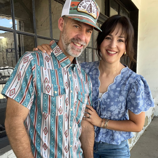 A smiling man and woman standing together outdoors in front of a building with large windows. The man wears a teal, white, and red patterned short-sleeve button-up shirt with blue jeans and a white cap, while the woman wears a blue floral puff-sleeve top and blue jeans.