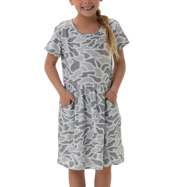 A young girl smiling and wearing a short-sleeve gray and white camouflage print dress with side pockets, standing against a plain white background.