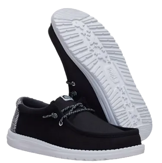 A pair of black slip-on shoes with white soles, shown against a light cream background. One shoe is positioned upright to display the textured white outsole, while the other lies flat to show the black upper and decorative grey laces.