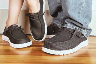 A close-up lifestyle shot of a person's legs and feet wearing a pair of dark brown slip-on knit shoes with white soles, standing on a wooden floor next to another person in jeans.
